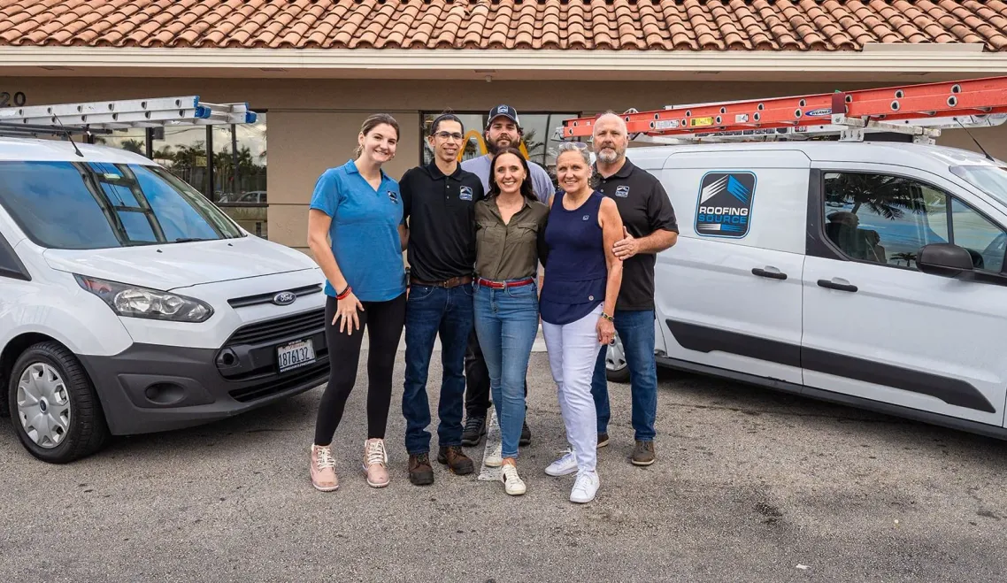 Six RoofingSource teammates standing together between two ladder-racked company vans outside a client restaurant.
