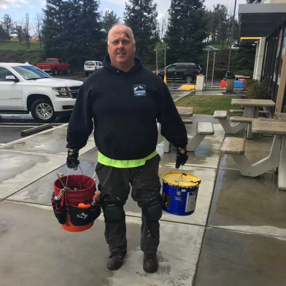 A teammate in a black RoofingSource hoodie carrying two buckets across a wet restaurant lot.