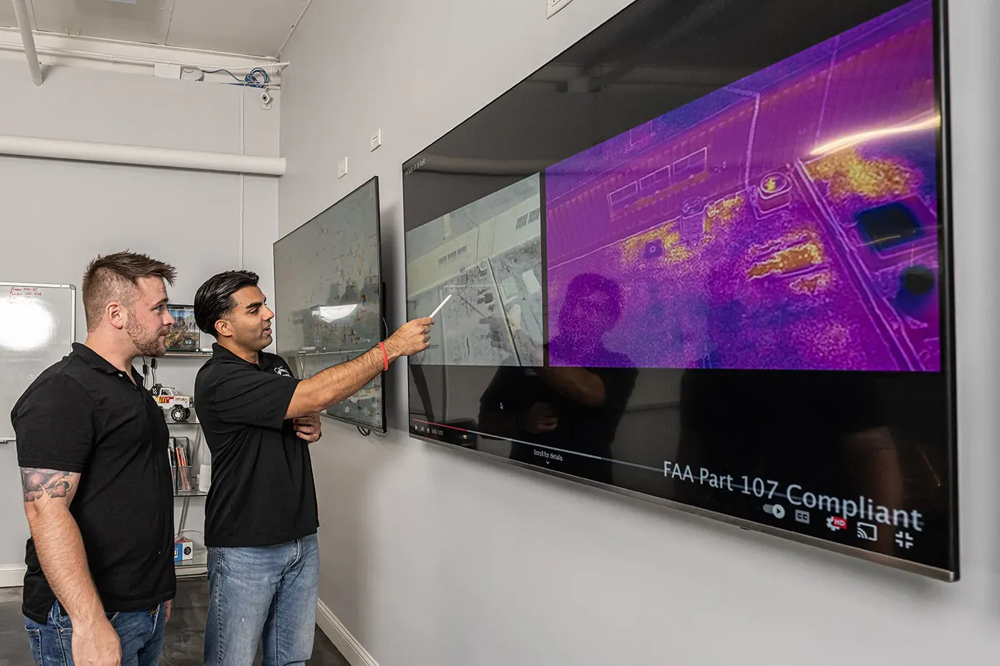 Two teammates studying a thermal aerial scan of a rooftop on a wall-mounted screen.