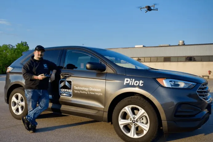A RoofingSource teammate with a drone controller beside a company car, the drone in the air above