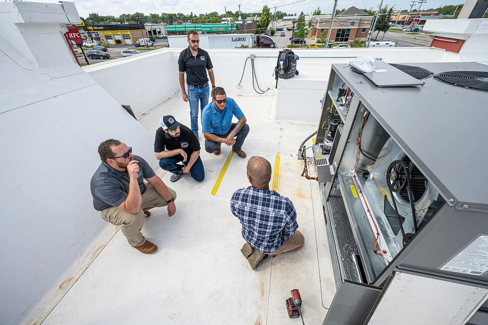 Five teammates around a trainer kneeling at an open rooftop unit on the HQ training deck