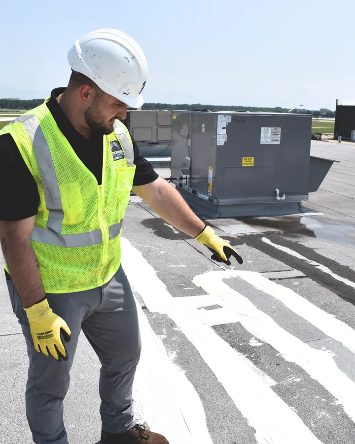 A teammate in a hard hat and safety vest looking over a roof membrane carrying several old sealant repairs