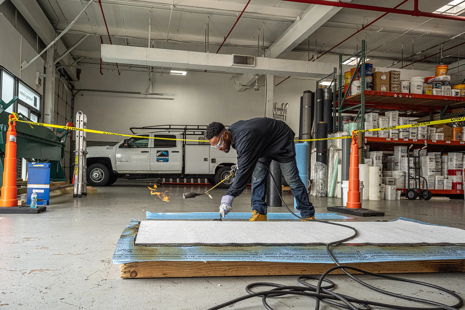 A teammate torching a membrane seam on a mock deck in the vehicle bay, a RoofingSource truck and racks of roofing material behind him.