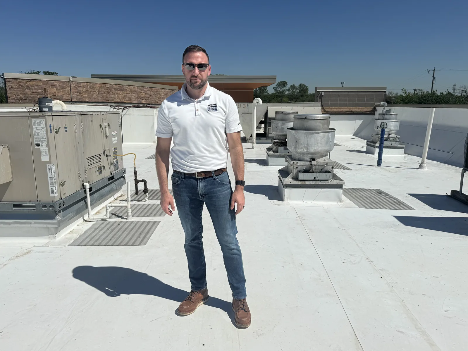 A RoofingSource teammate in a white company polo standing on a client’s white rooftop among the exhaust and air-handling units