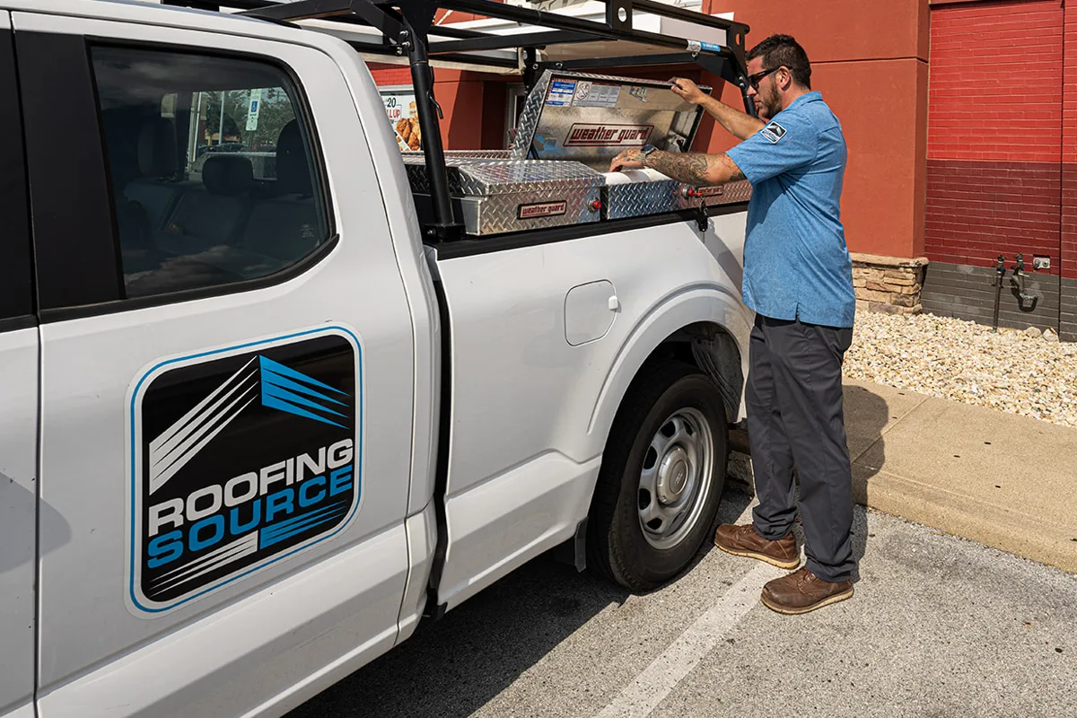 A RoofingSource Field Advisor reaching into the open toolbox of a company truck at a client site, the RoofingSource mark on the door