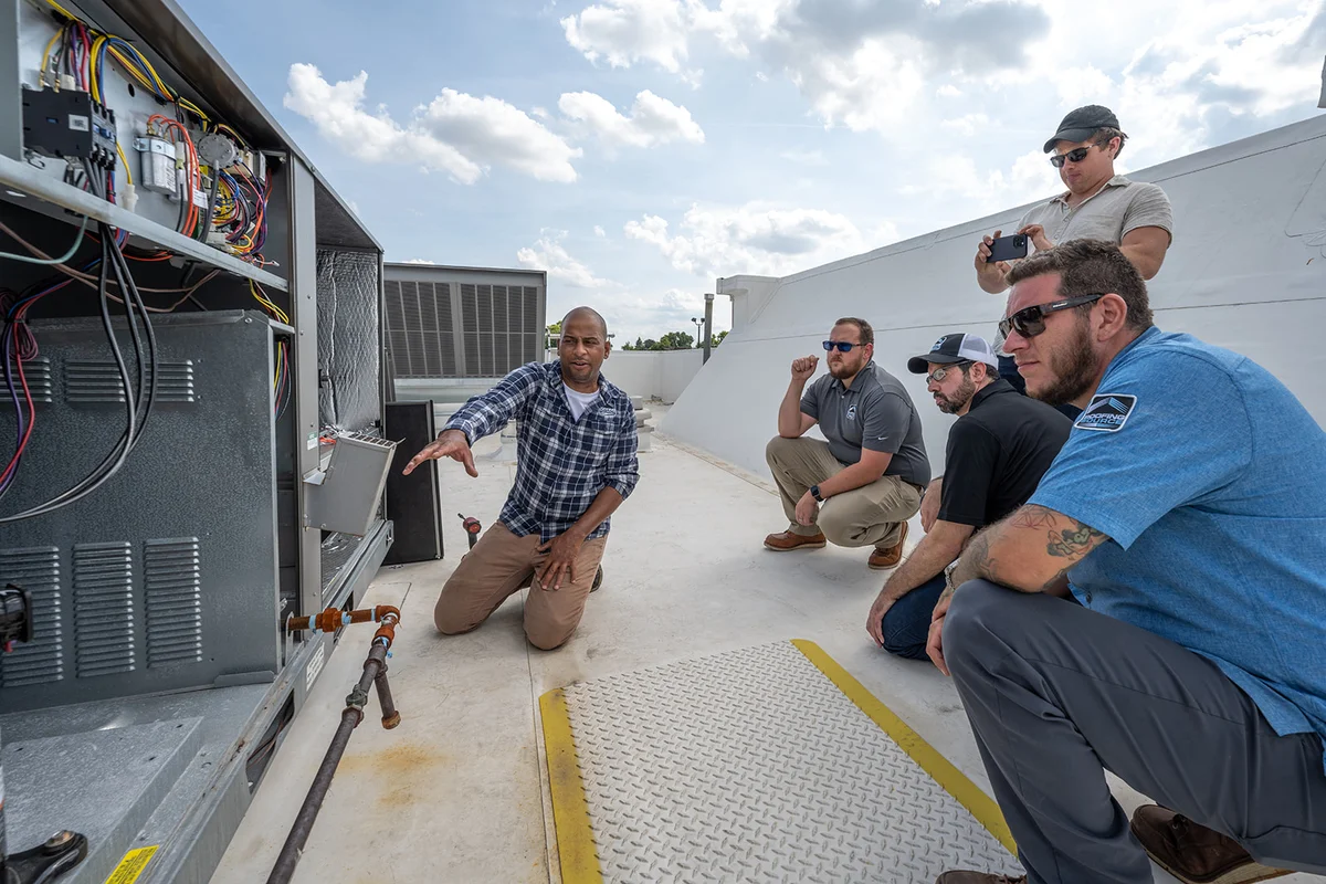 A trainer kneeling at an open rooftop unit on the HQ training deck, four teammates listening