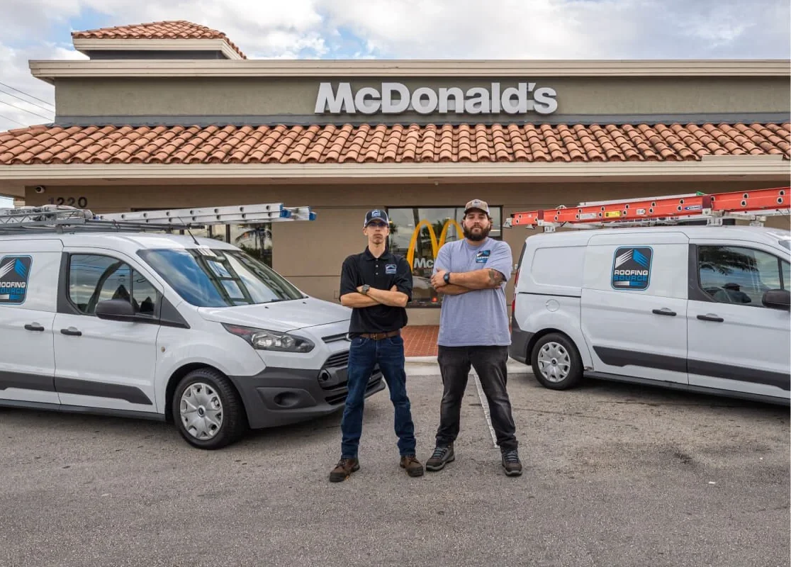 Dos compañeros de RoofingSource junto a dos furgonetas de la empresa con escaleras en el techo, frente a un restaurante cliente con techo de tejas y palmeras
